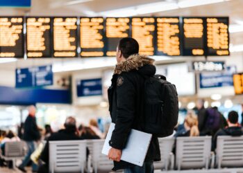 man standing inside airport looking at LED flight schedule bulletin board