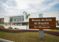 Fotografia da fachada contemporânea do Museu de Arte de Brasília com placa de sinalização bilíngue (português e inglês), vegetação paisagística e céu parcialmente nublado ao fundo.