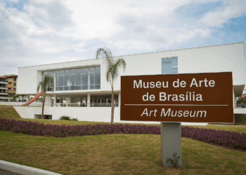 Fotografia da fachada contemporânea do Museu de Arte de Brasília com placa de sinalização bilíngue (português e inglês), vegetação paisagística e céu parcialmente nublado ao fundo.