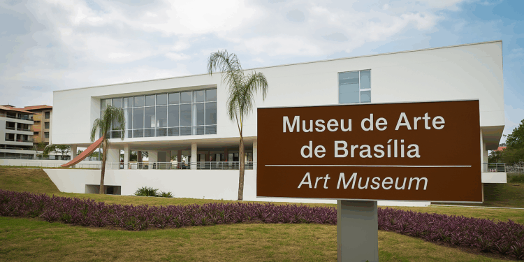 Fotografia da fachada contemporânea do Museu de Arte de Brasília com placa de sinalização bilíngue (português e inglês), vegetação paisagística e céu parcialmente nublado ao fundo.