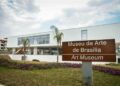 Fotografia da fachada contemporânea do Museu de Arte de Brasília com placa de sinalização bilíngue (português e inglês), vegetação paisagística e céu parcialmente nublado ao fundo.